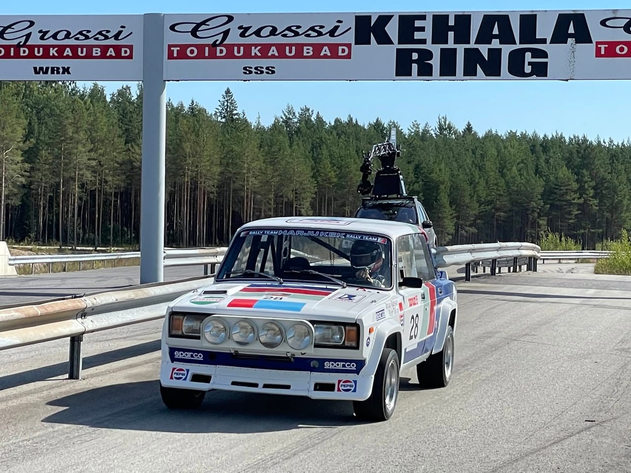 The restored Lada, competition number 28 in red, white and blue livery with a four-lamp pod and Sparco and Pepsi decals, running on asphalt under a gantry lettered KEHALA RING. A HARJU KEK RALLY TEAM band crosses the top of the windscreen and a helmeted driver is at the wheel. Close behind and to the right, a light-coloured 4x4 carries a cinema camera on a roof-mounted rig. Pine forest and guardrails on both sides under a clear sky.