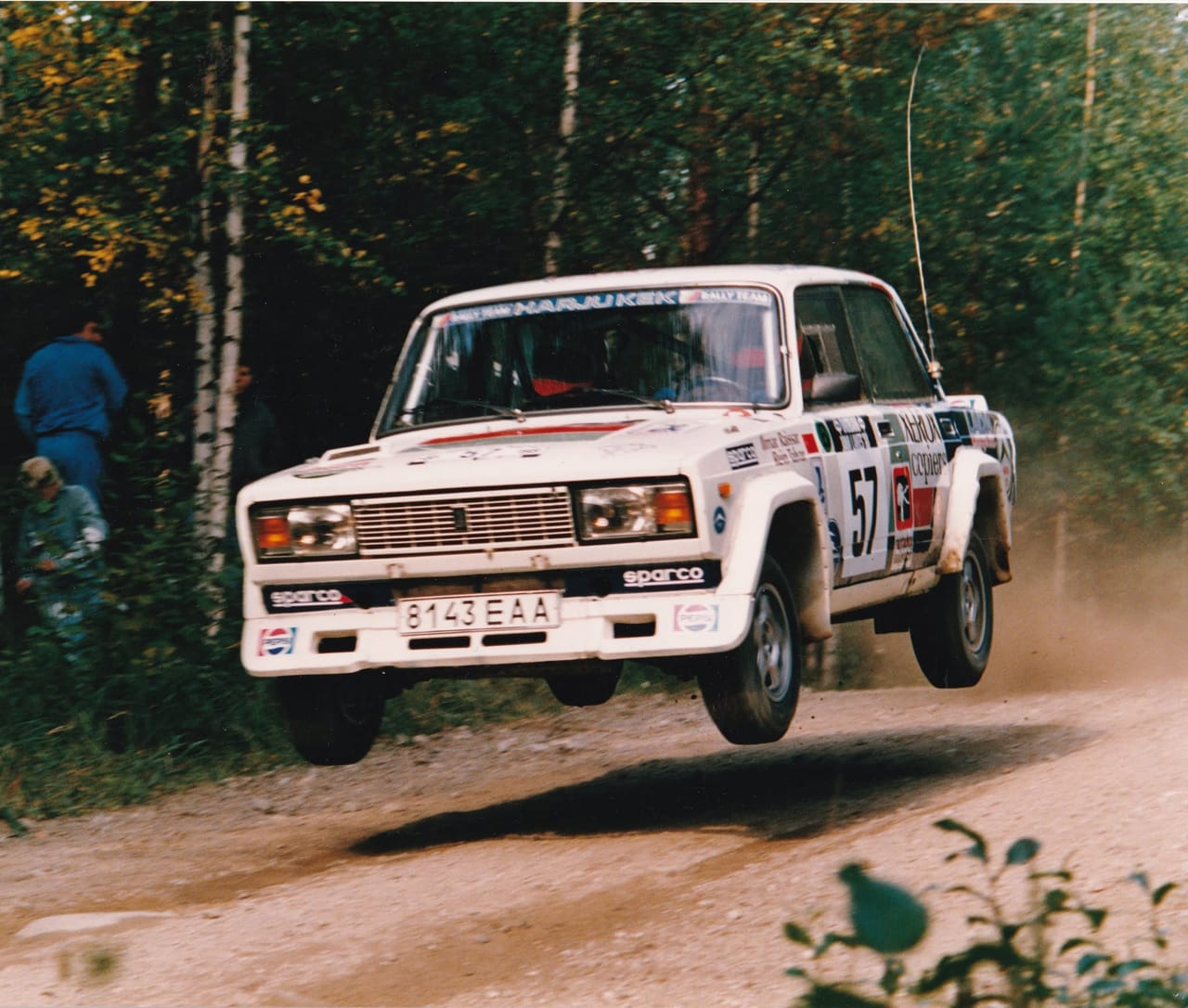 A white Lada saloon in Harju KEK Rally Team livery, competition number 57, fully airborne over a gravel crest in autumn birch forest. The front wing carries the names Ilmar Raissar and Rein Talvar; the plate reads 8143 EAA, with Pepsi and Sparco decals along the bumper. Two spectators, an adult and a child, stand among the trees on the left.