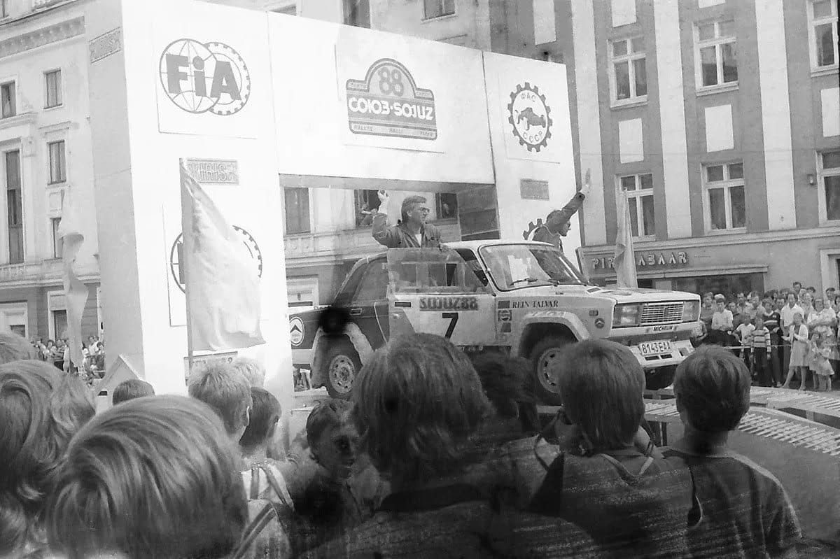 Black and white photograph of a rally start ramp in a city street. The arch above it carries three banners: the FIA emblem on the left, the SOIUZ-88 rally logo in the middle in Cyrillic and Latin, and the cogged emblem of the Soviet motor sport federation on the right. A Lada saloon numbered 7, lettered REIN TALVAR on the front wing, sits on the ramp with a crew member standing up through the roof and waving. A dense crowd of onlookers fills the foreground and lines the far pavement.